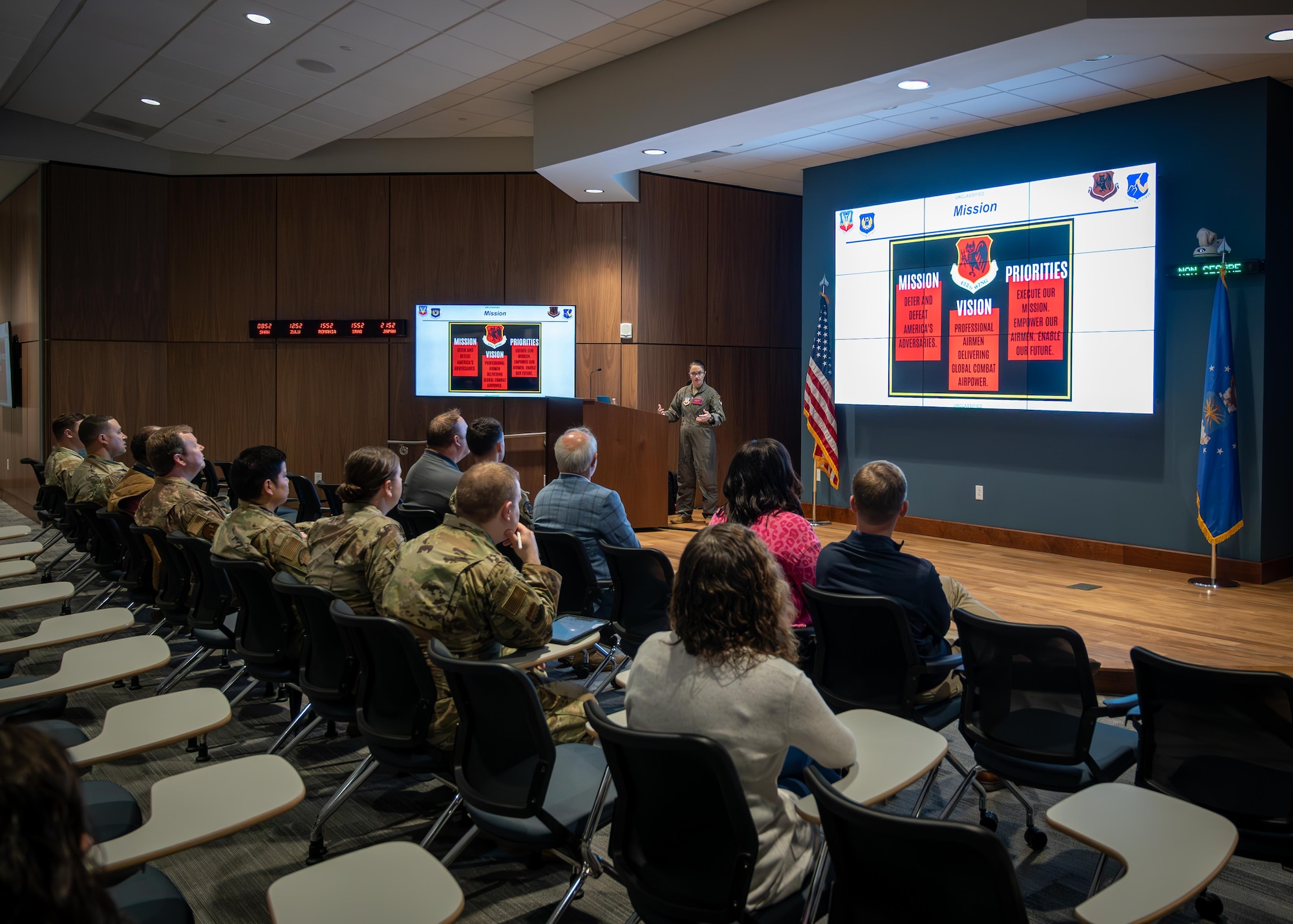 Civilians and military personnel listen to a brief in an auditorium.