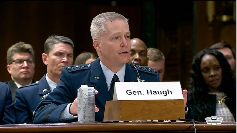 A man in military dress coat with four stars on each shoulder is sitting at a table with a microphone and small podium in front of him. An assortment of civilians sits in the background.