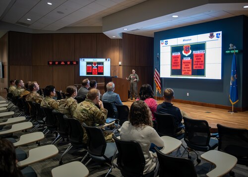 Civilians and military personnel listen to a brief in an auditorium.