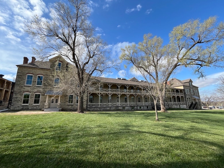 A two-story limestone building with two trees and grass in the foreground and a blue sky in the background.