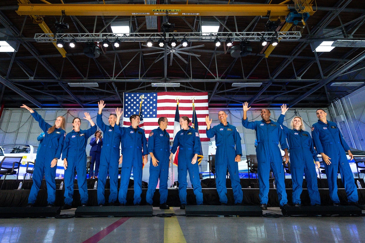 Ten people in flight suits wave in a warehouse. A large U.S. flag is displayed behind them.
