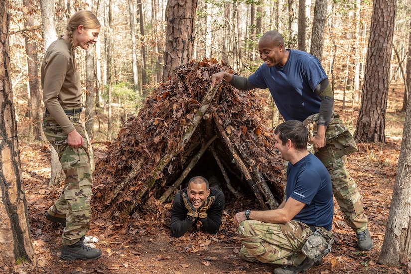 Four people admire a tent they made in the woods using sticks, leaves and tree branches.