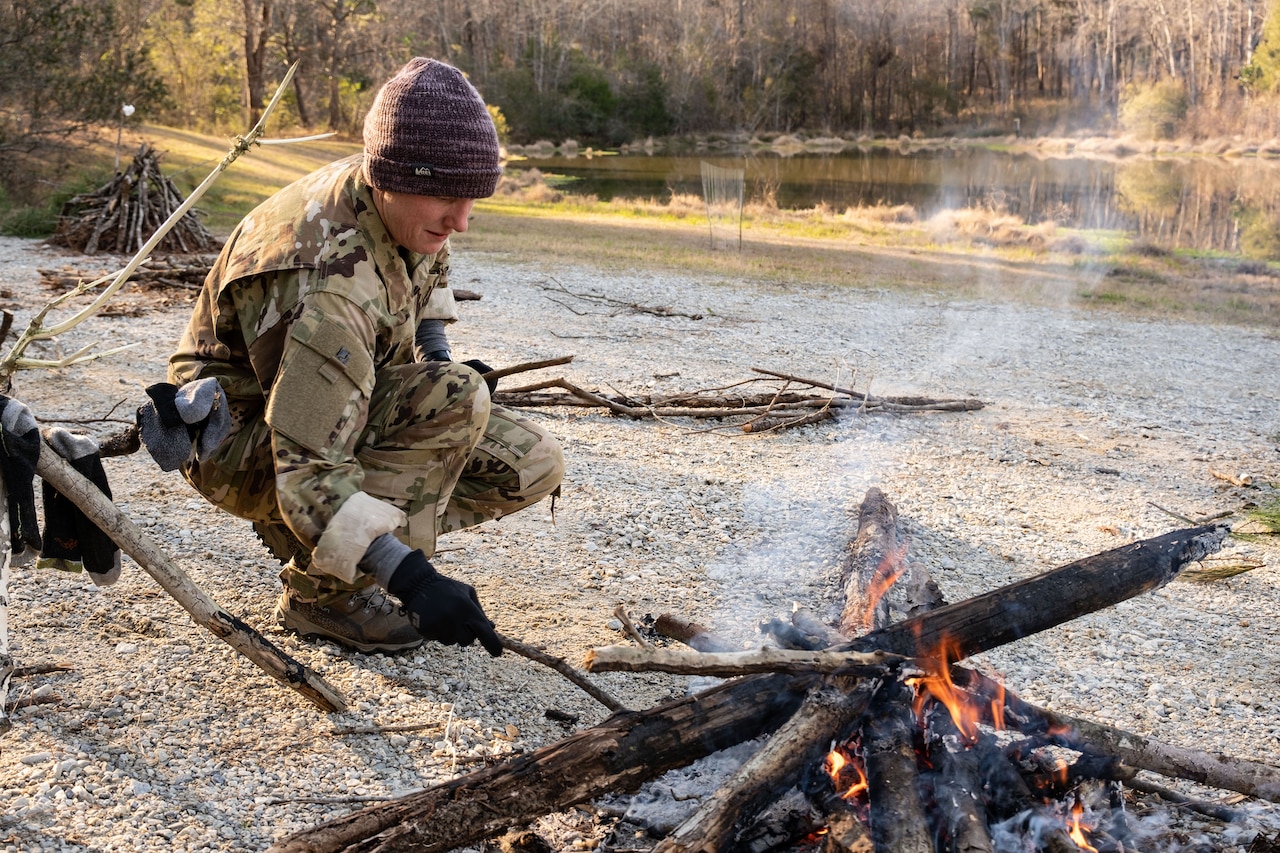 A person hunches over a small fire, poking it with a stick.