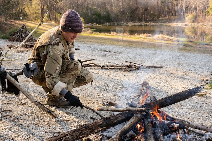 A person hunches over a small fire, poking it with a stick.