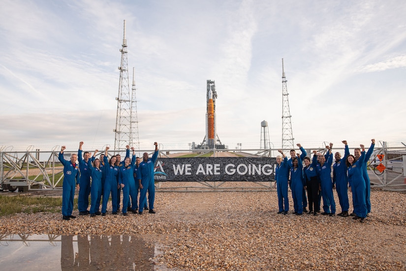 Nearly two-dozen people pose with their fists in the air in front of a gate. Behind the gate is a massive rocket launch platform.