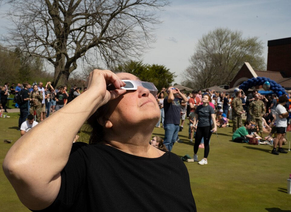 A woman in the foreground holds a pair of eclipse glasses to her eyes as she looks skyward. Behind her, other people talk and look up.