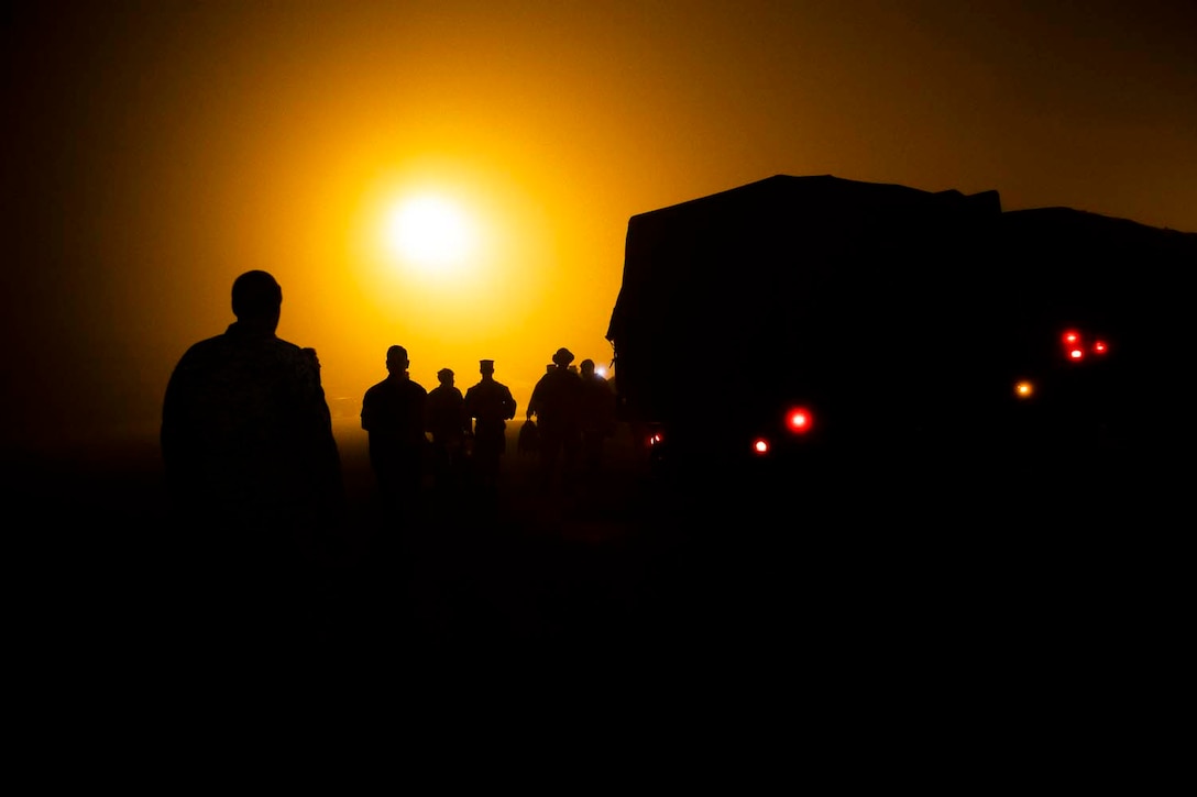 Marines walking toward a large, darkened vehicle are silhouetted against the sun in an orange sky.