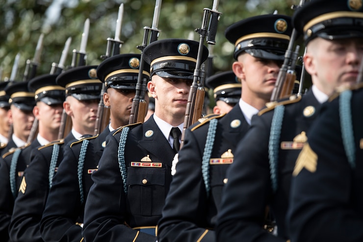 Army Soldiers dressed in dark ceremonial uniforms are standing in a row during a ceremony while carrying rifles with bayonets affixed.
