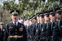 Army Soldiers in dark ceremonial unforms with rifles are standing in a row while one member standing to the side is calling out orders while carrying a sword.