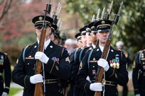 Army Soldiers stand in formation rows dressed in dark ceremonial uniforms and white gloves while holding brown rifles in front of them with both hand.