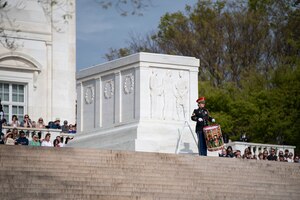 An Army Soldier is playing a large field drum while standing at the top of steps next to the white marble clad Tomb of the Unknown Soldier. There are people in the background watching the ceremony.