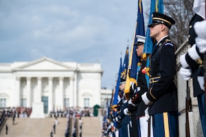 Dozens of service members from each branch of the military are holding flags and rifles while lining the walkway leading up to the Tomb of the Unknown Soldier and the white-columned building behind it.