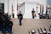 An Army band drummer is performing on a large field drum at the top of the steps leading up to the Tomb of the Unknown Soldier while dozens of military members from all branches are marching up to line the sides of the steps for a wreath ceremony.