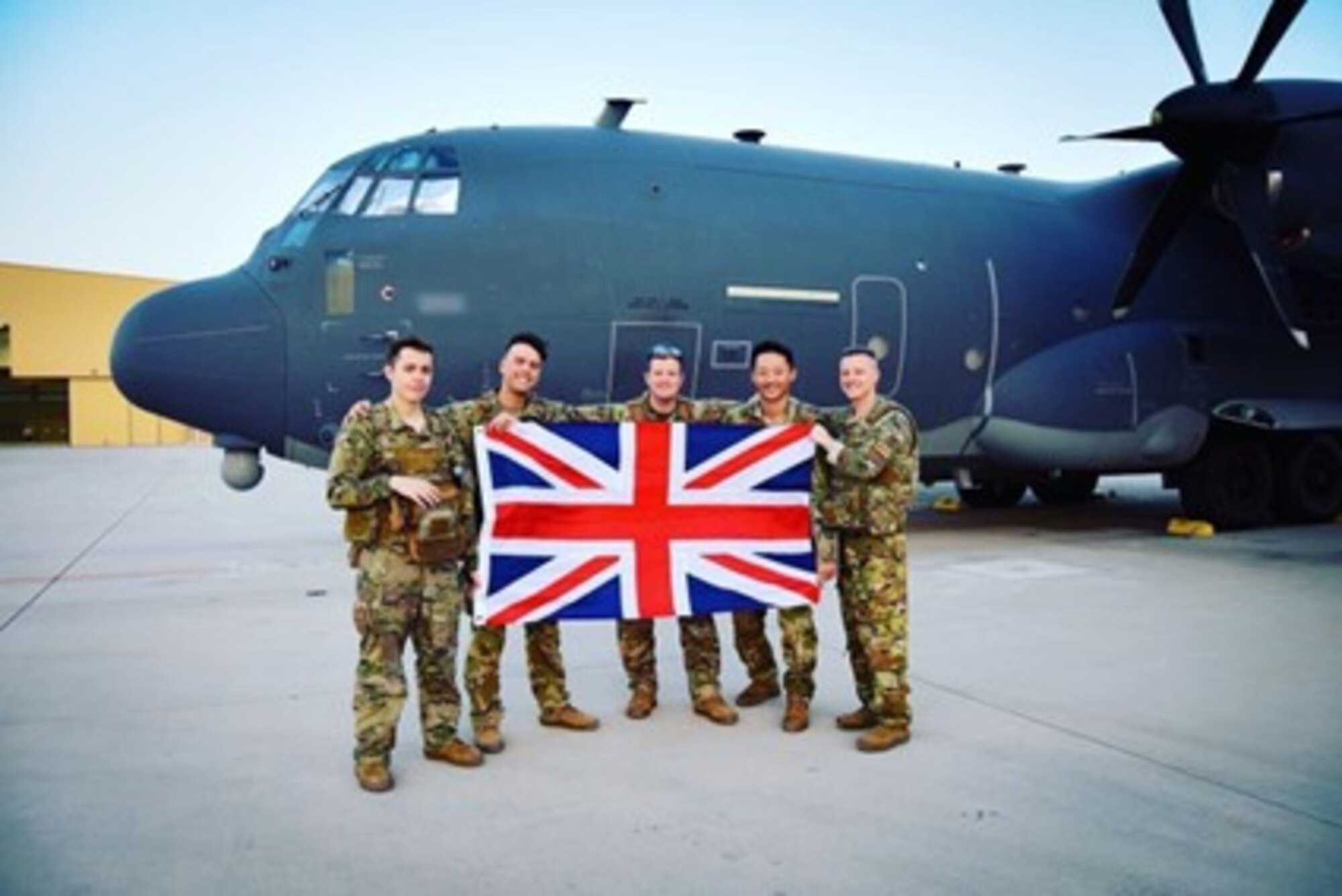 A group of five individuals in military uniform pose with a U.K. flag in front of a military aircraft.