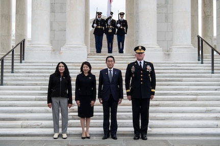 Four people are posing for a picture on white marble steps. At the top of the steps, two Army Soldiers are holding rifles on either side of another Soldier who is holding the Japanese flag.
