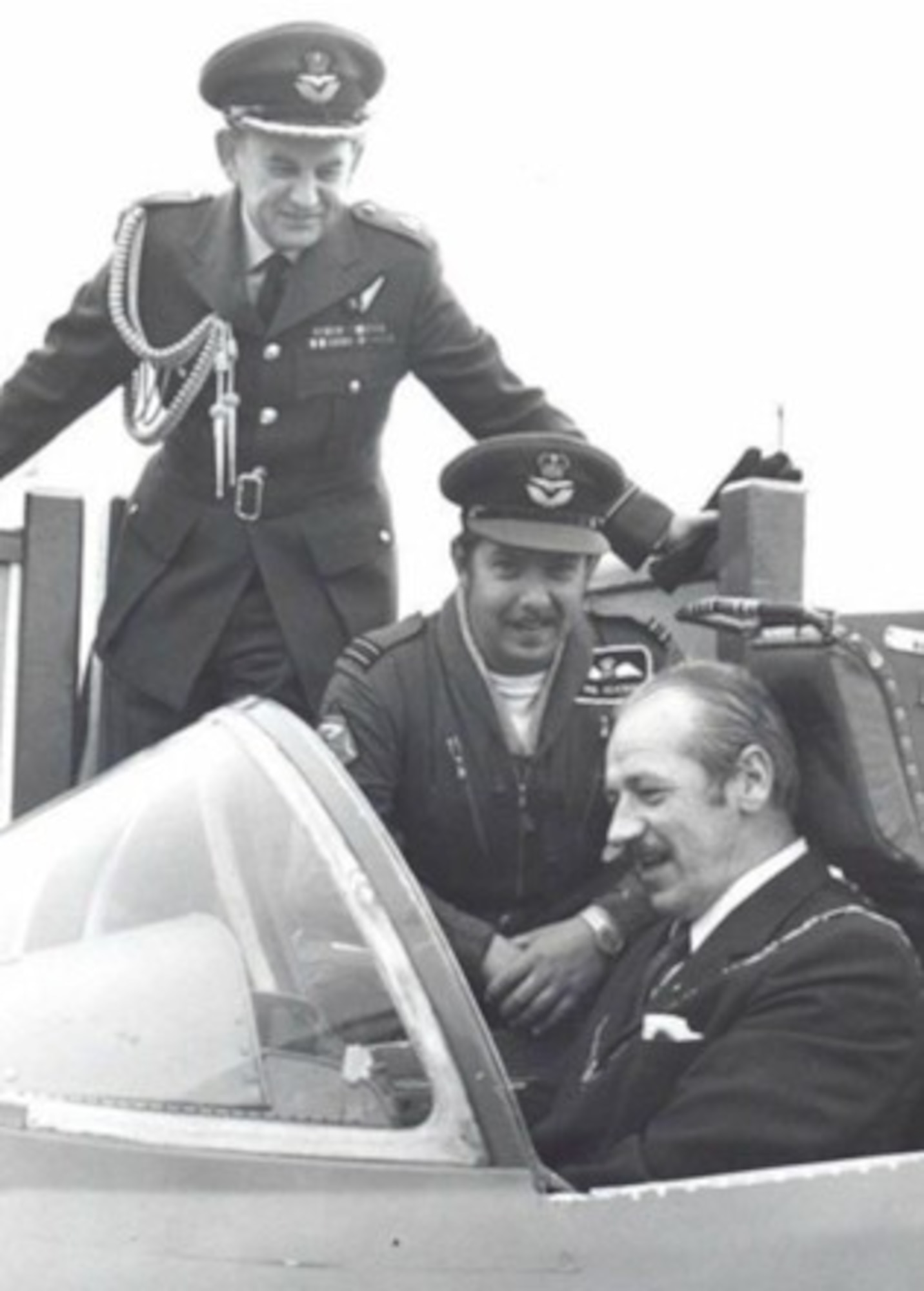 A man sits in a military aircraft cockpit while two other men in military uniform look on.