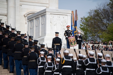 An Army band drummer is dressed in ceremonial uniform playing on a large snare drum at the top of stairs. Ceremonial honor guardsmen from various branches of the military are marching up the steps on either side of the drummer.