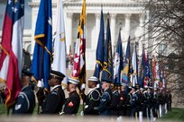 Military members from various branches of the military are lined up on a walkway holding various flags.