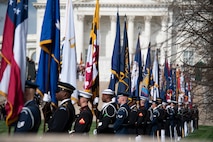 Military members from various branches of the military are lined up on a walkway holding various flags.