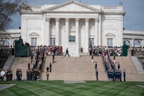View of the steps up to the back side of the white marble Tomb of the Unknown Soldier and the building behind it that houses the history museum for the Tomb. There are various military members from each branch lining the steps.