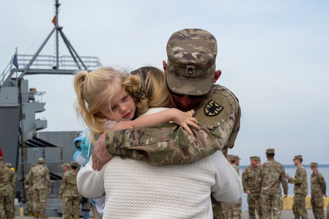 A soldier hugs his wife and daughter at a pier with a vessel and other soldiers in the background.