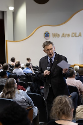 Man in suit holding microphone in one hand and paper in the other, speaks to audience.