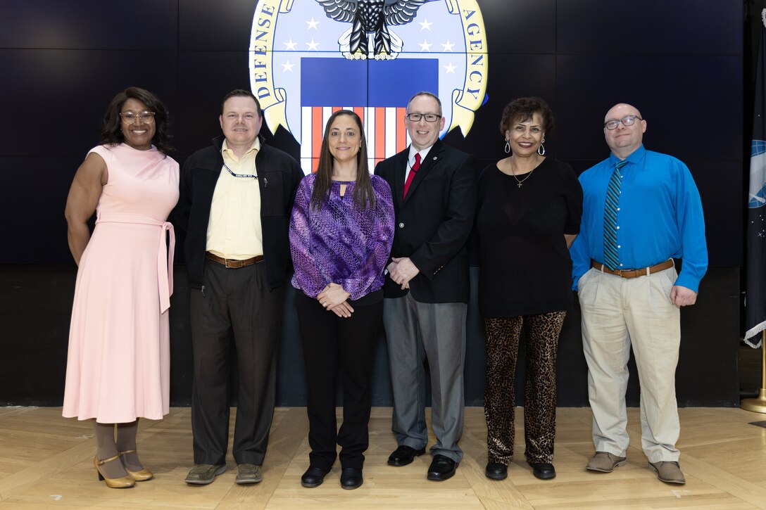 Six people pose for photo , wood floor, black background with DLA logo