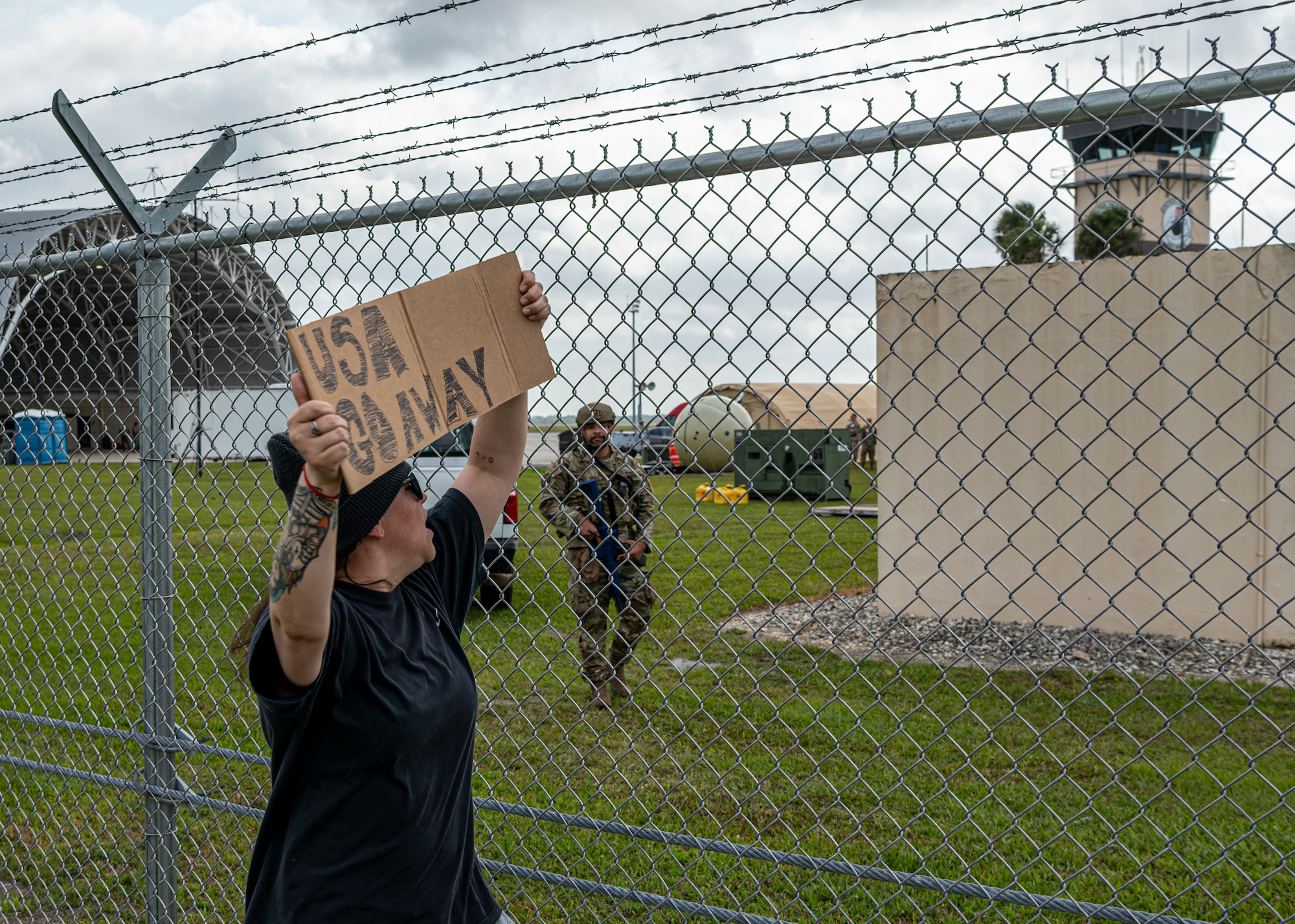 A person wearing all black clothes holds a sign, "USA GO AWAY" outside of a gate, simulating protest.