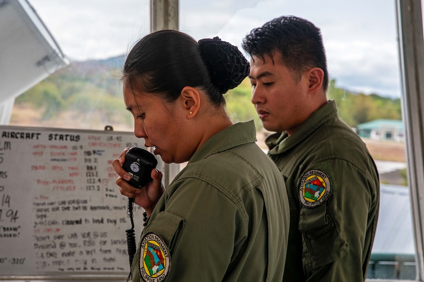 Two service members stand in front of a whiteboard with text in an enclosed space with one talking on a communications radio.