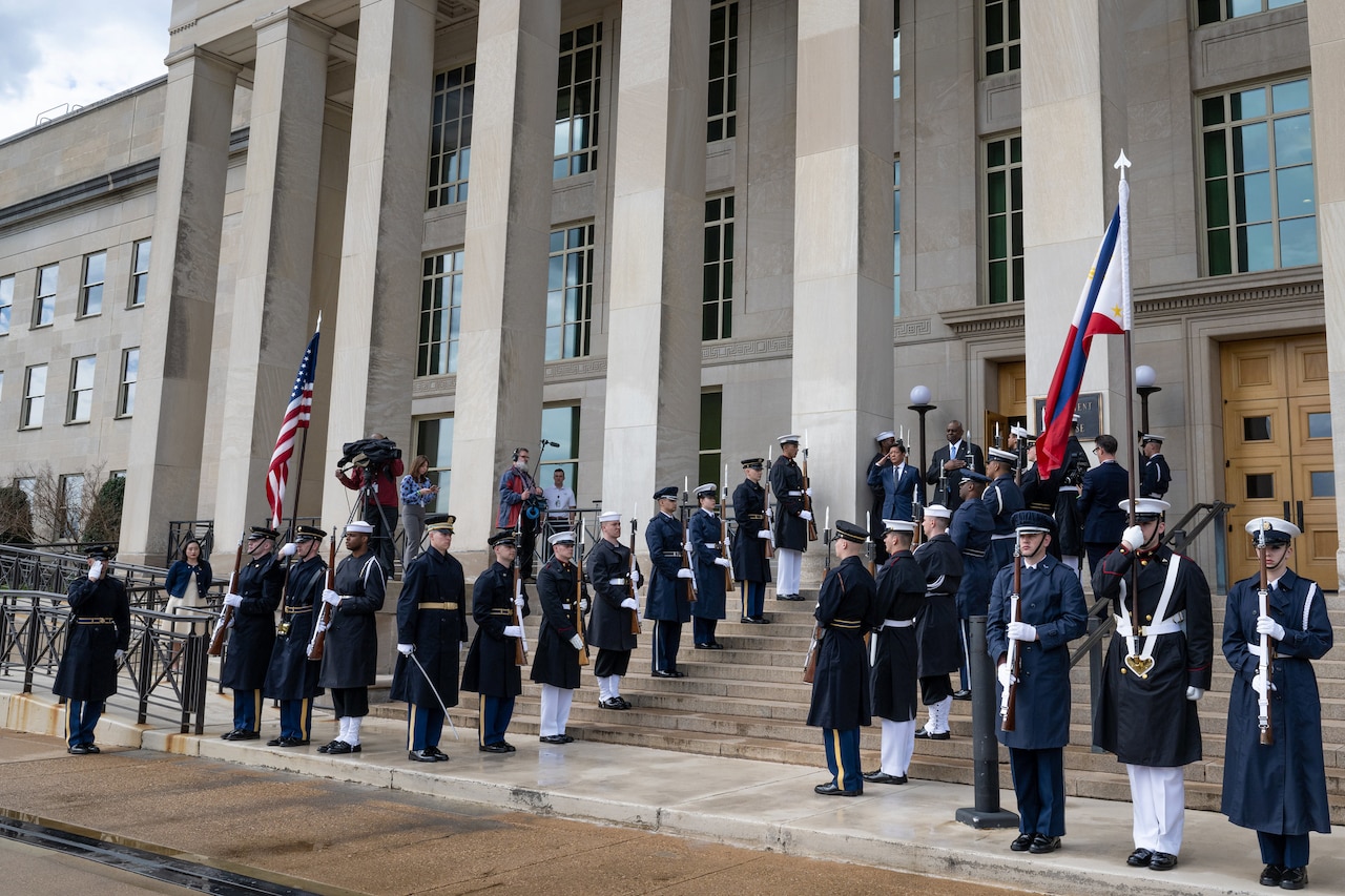 Secretary of Defense Lloyd J. Austin III stands with another official atop steps outside the Pentagon, flanked by honor guard members.