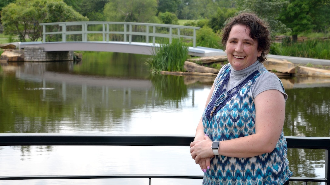 Woman wearing blue top stands along a railing with water and a bridge in the background.