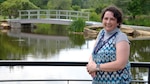 Woman wearing blue top stands along a railing with water and a bridge in the background.