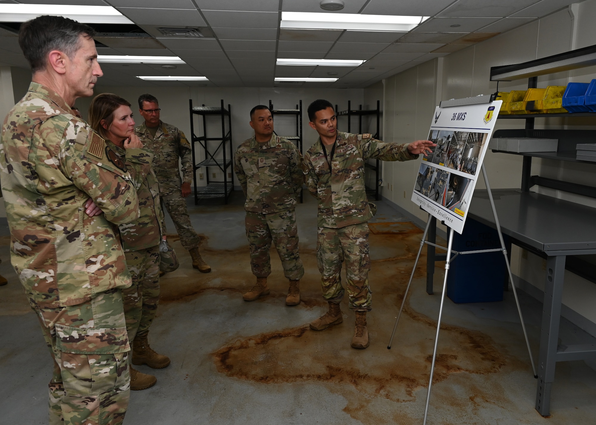 Four Airmen looks at a board that the fifth Airman points to. The board contains photos of the damage Typhoon Mawarr caused.
