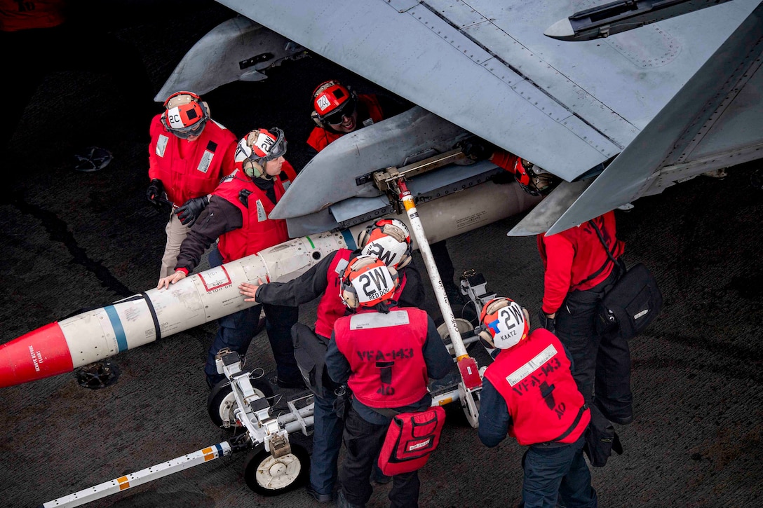 Overhead shot of sailors loading ordnance onto an aircraft from the flight deck of a Navy ship.
