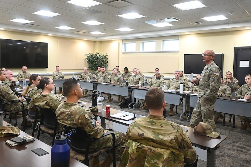Uniformed service members are seated around a conference table as a standing service member speaks.