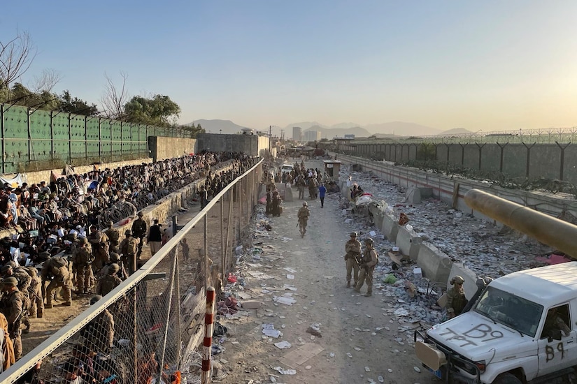 Over a dozen service members in camouflage and hundreds of evacuees are on one side of a chain link fence while a handful of other camouflaged service members patrol on the opposite side of the fence.