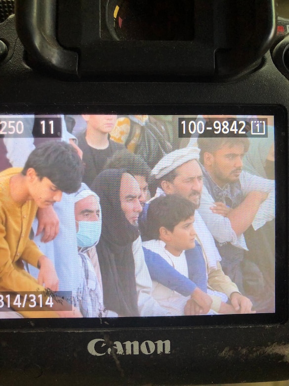 An image of a bald man in a black shirt and matching black head scarf who is sitting surrounded by Afghan refugees. The image is pulled up on the back of a still photo camera.