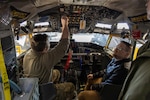 A man in a tan camouflage uniform sits in a airplane cockpit seat and reaches up to touch some of the controls to show another man in business casual clothing some of the systems in a KC-135 cockpit. The man in casual business attire has glasses and a beard and is looking up.