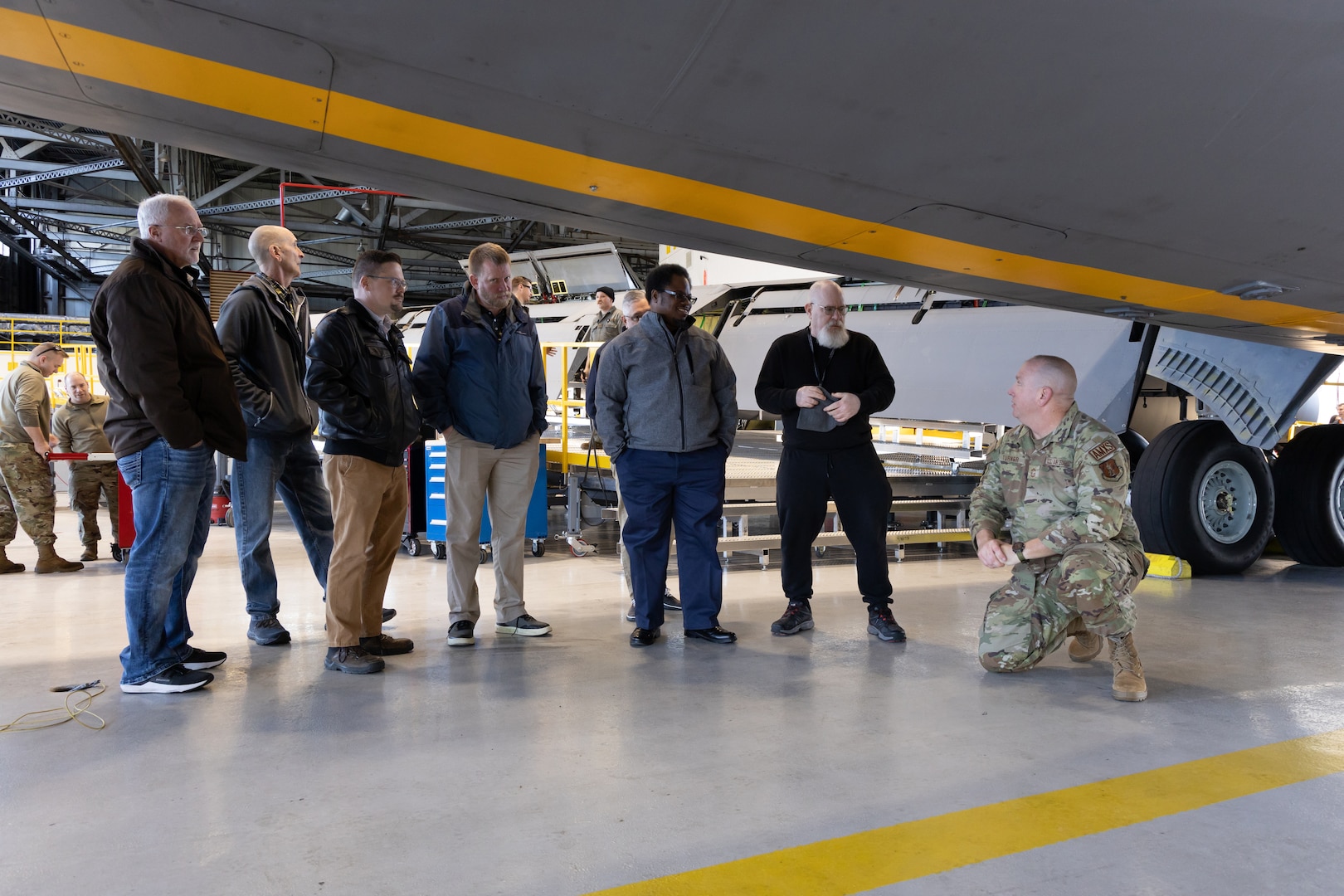 A bald man in a camoflage uniform squats underneath a gray cargo/refueling plane undergoing phase maintenance and inspection or ISO. It is surrounded by scaffolding and is inside a hanger. He is speaking to a group of men in business casual attire.
