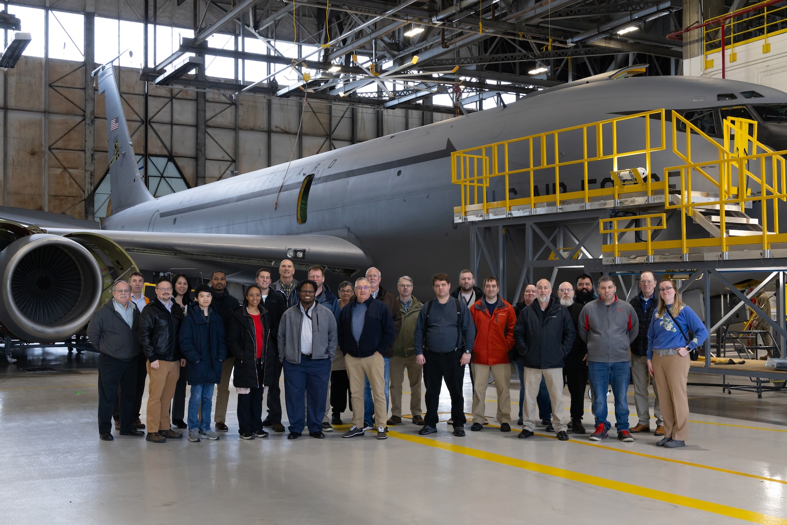 A large group of people stand in front of a gray cargo/refueling plane undergoing phase maintenance and inspection or ISO. It is surrounded by scaffolding and is inside a hanger.