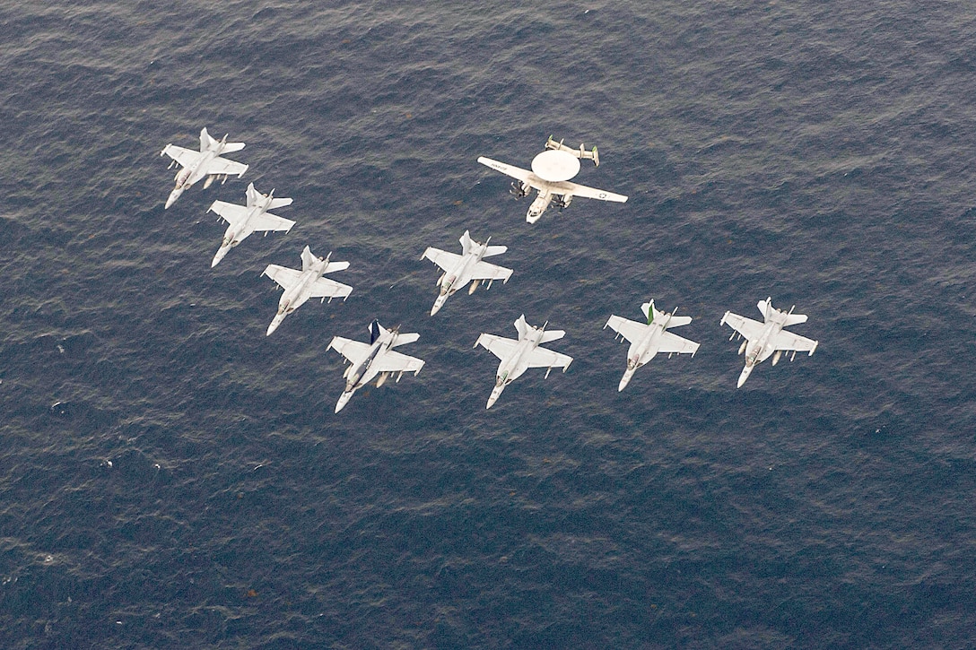 Eight aircraft fly in a diagonal arrow formation to the bottom left over a body of water.