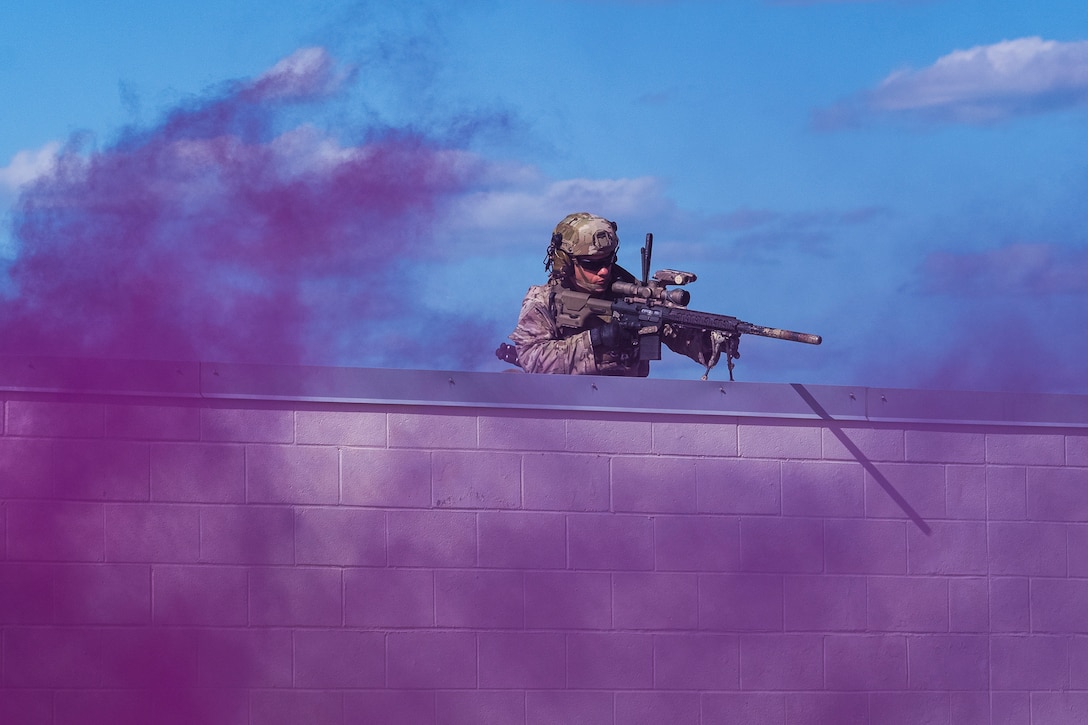 A soldier stands behind a white structure while holding a weapon as purple clouds of smoke fill the air.