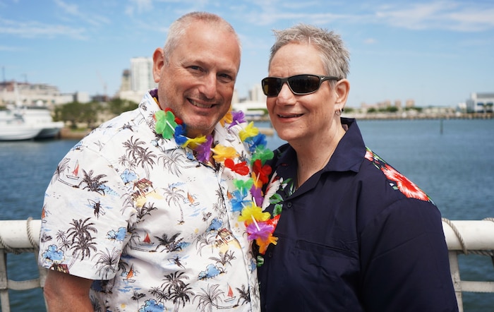 Robert Kopser on the left in a Hawaiian themed button-up shirt with his wife Jennifer also in a button up shirt but her's is a dark navy color. They are in front of the water aboard a boat.