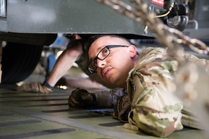 An Airman in uniform lays on the floor of a C-5M Super Galaxy and peers under a vehicle.