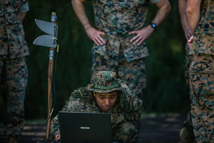A Marine works on a laptop on the ground outdoors as three others stand by him.