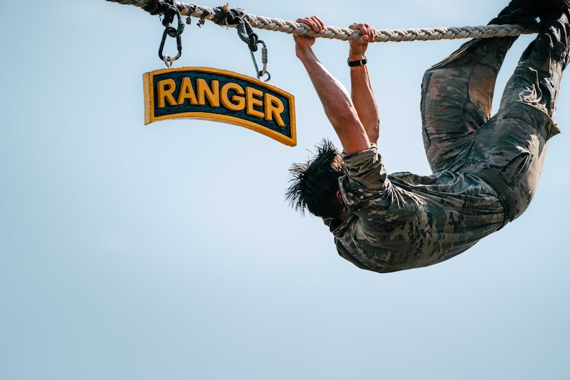 A soldier uses arms and legs to hang upside down from a rope. A sign to the soldier's left says ‘Ranger.’