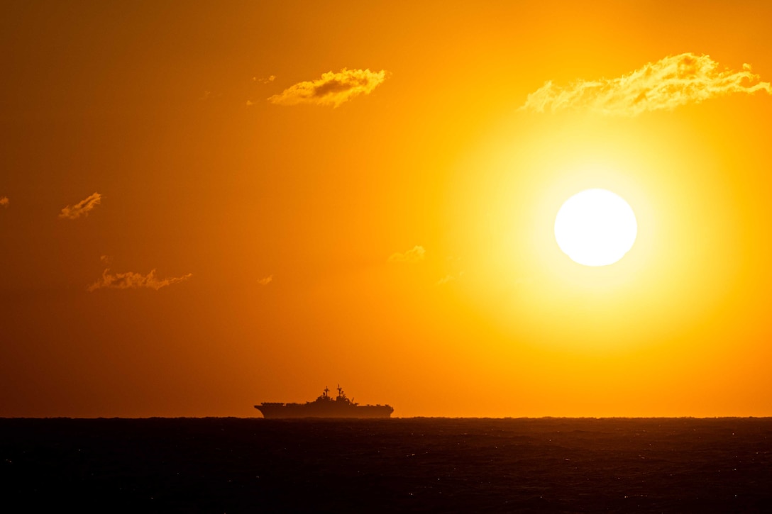 The sun rises into an orange sky as a military ship moves across the ocean.