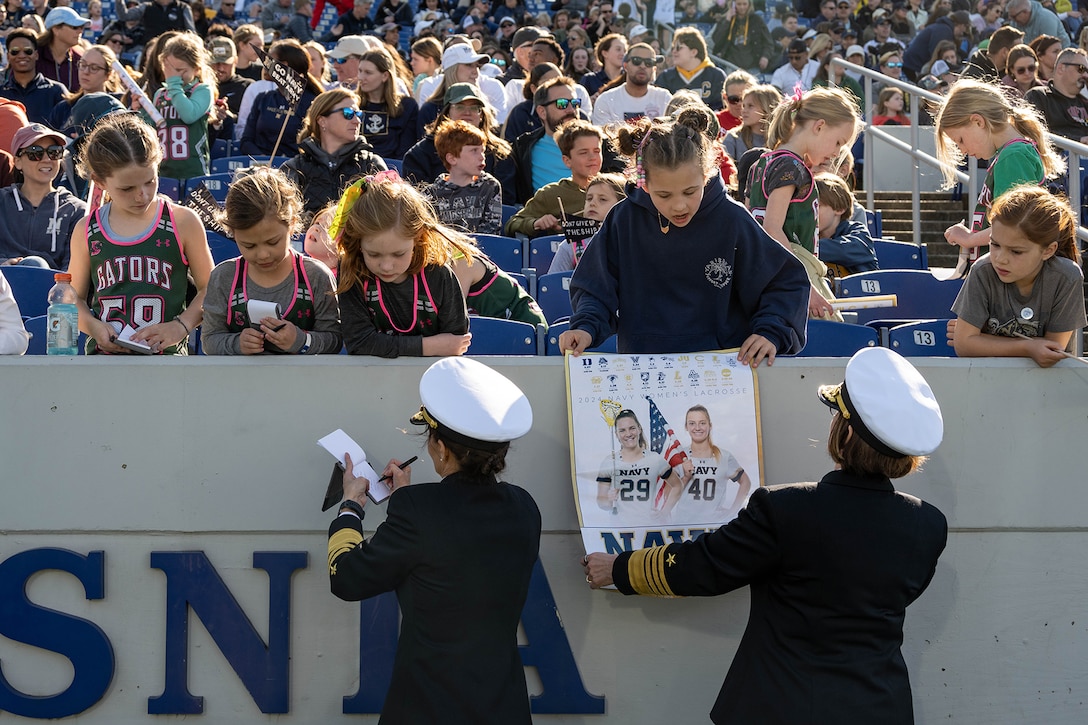 Uniformed service members sign autographs and greet young fans at a lacrosse game.