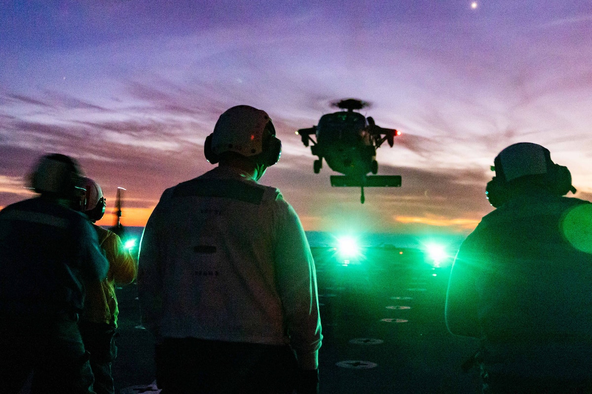Sailors wearing ear protection await the landing of a military helicopter on the flight deck of a Navy ship at twilight. Green lights can be seen on the flight deck.