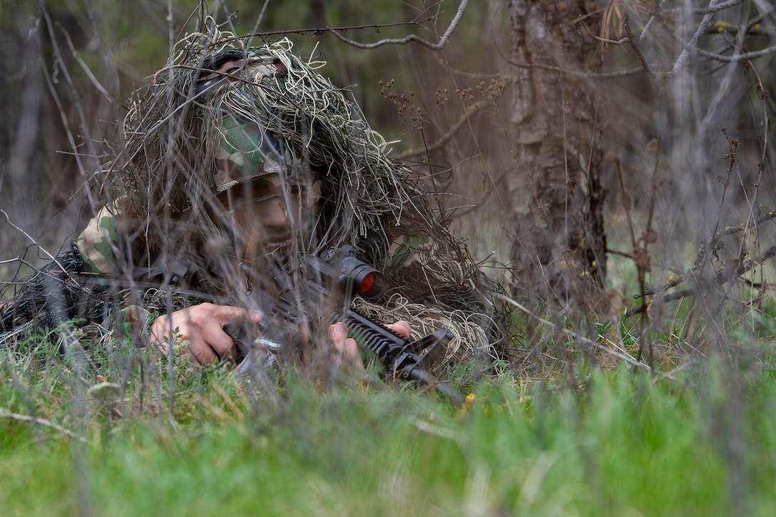 A service member holding a weapon in a wooded area lays under grass-like camouflage during training.